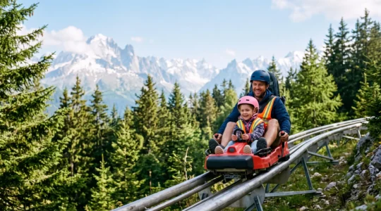 Un parent et son enfant dévalent ensemble une piste de luge d'été sur rail dans un paysage alpin français, sous un ciel dégagé.